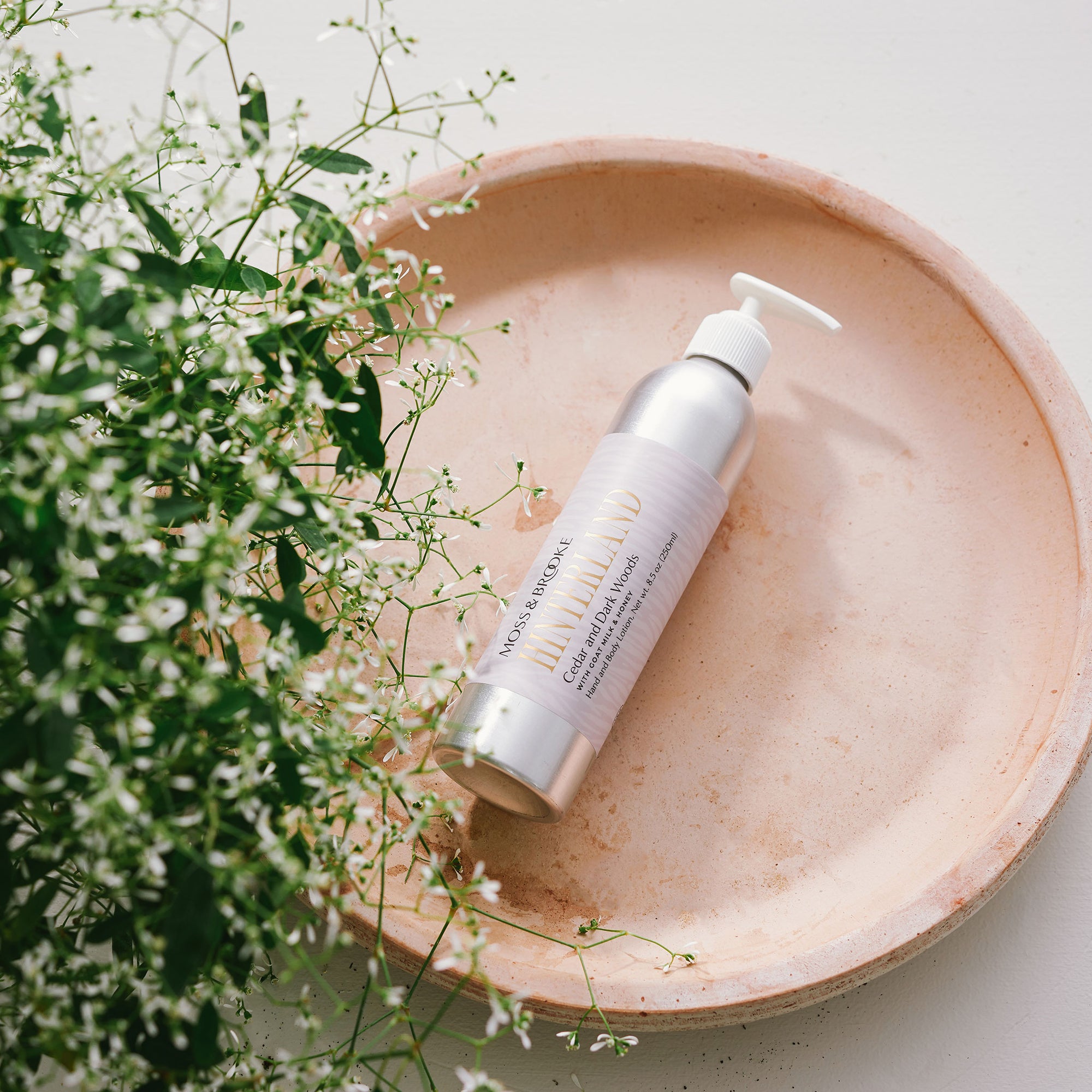 Bottle of Hinterland Body lotion by Moss & Brooke with flowers and greenery in background. Aluminum silver bottle with white pump and light grey label with gold text, sophisticated and luxurious.