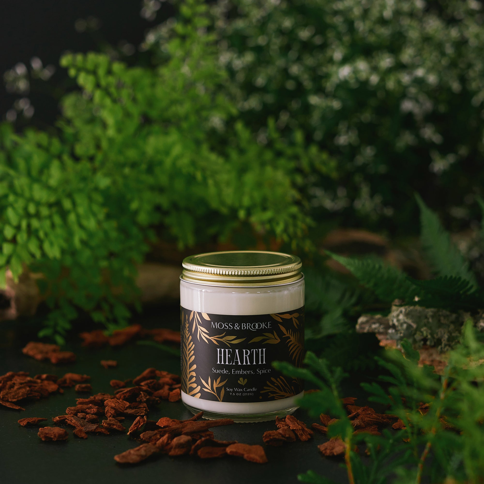Clear glass candle jar with gold lid and black label with metallic gold illustrations of leaves and ferns. Photo background with leaves, flowers and ferns. Hearth candle scented with notes of suede, smoky embers, cinnamon and spice.